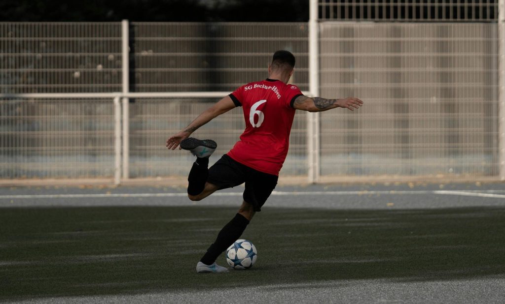 Un jugador de fútbol masculino con camiseta roja patea un balón en un campo al aire libre durante el día.