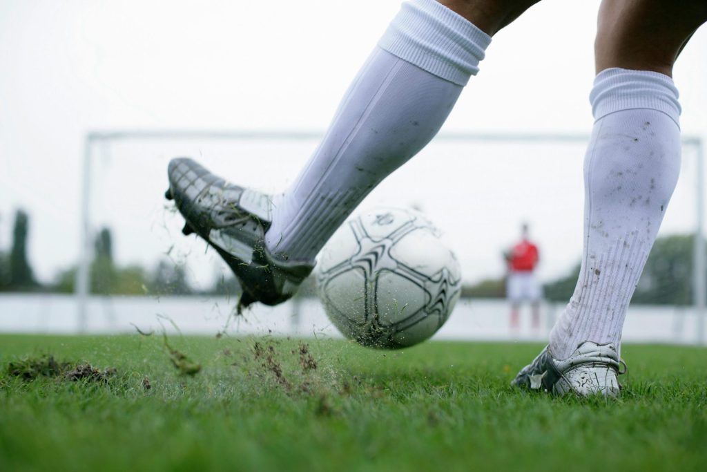 Primer plano de un jugador de fútbol pateando un balón con movimiento e intensidad en un campo de césped, mostrando acción y deportividad.