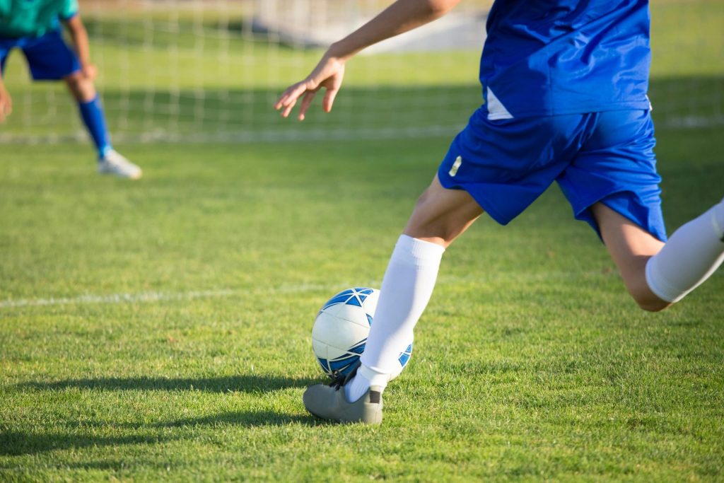 Un joven futbolista con uniforme azul patea un balón hacia la portería en un día soleado.
